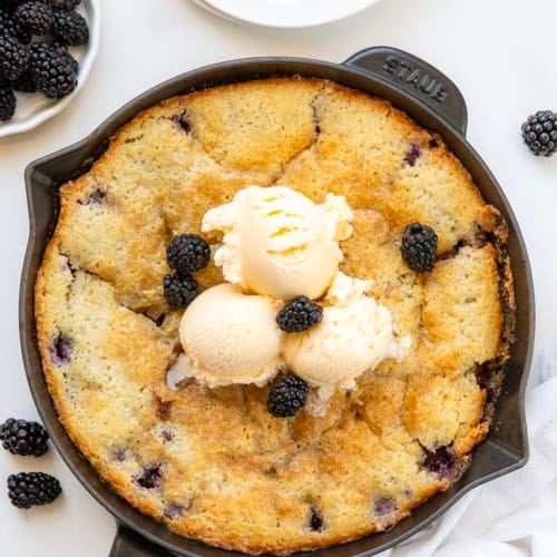 Skillet of Blackberry Cobbler on a white table with fresh blackberries from overhead.