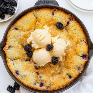 Skillet of Blackberry Cobbler on a white table with fresh blackberries from overhead.
