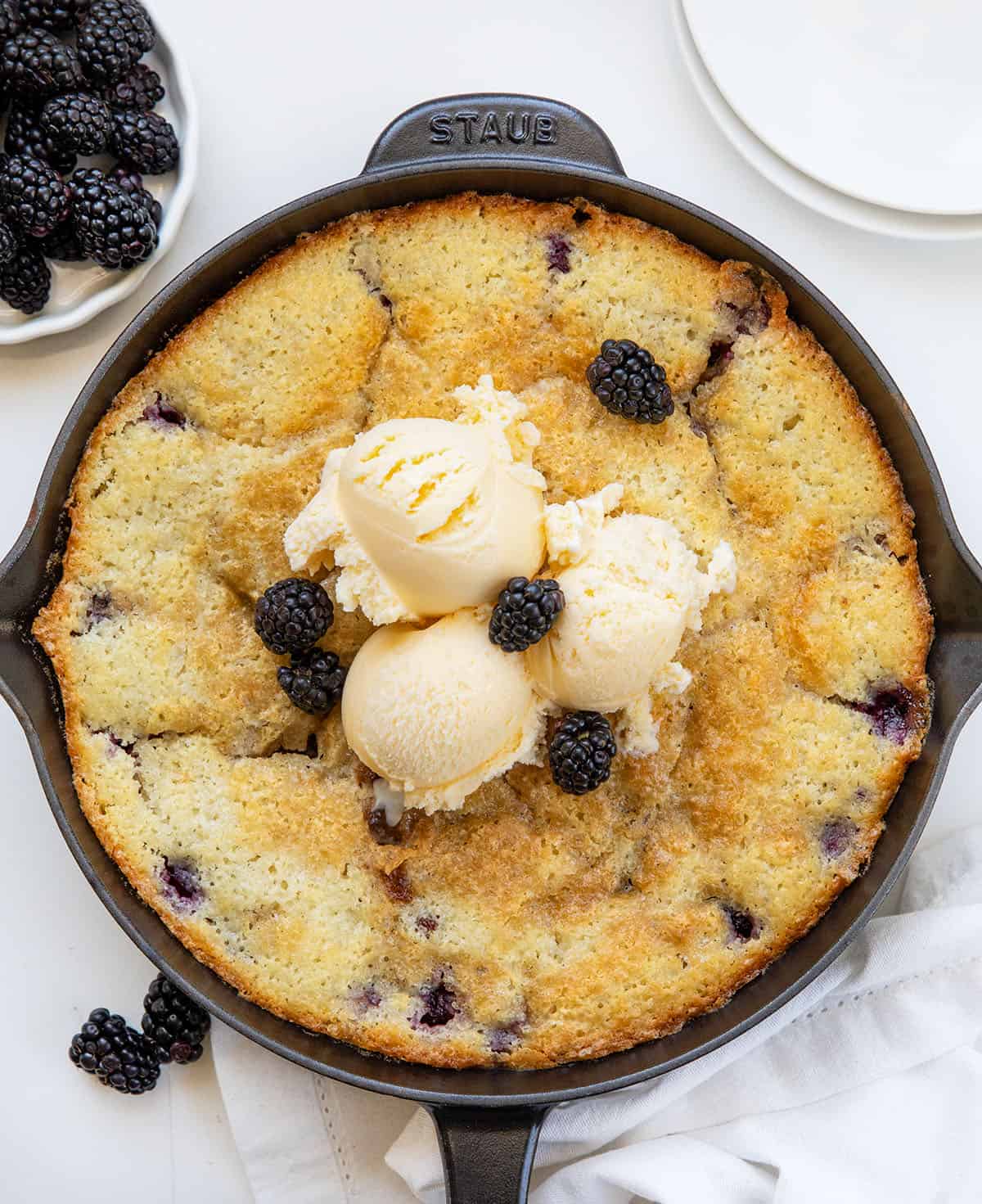 Skillet of Blackberry Cobbler on a white table with fresh blackberries from overhead.