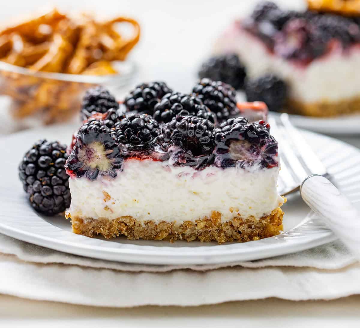 This Blackberry Pretzel Salad is a sweet and salty layered dessert made with a crunchy pretzel crust, creamy cheesecake-style filling, and a glossy black cherry gelatin layer packed with juicy blackberries. It’s creamy, fruity, and perfectly refreshing! Piece of Blackberry Pretzel Salad on a white plate on a white table with a fork resting on the plate.