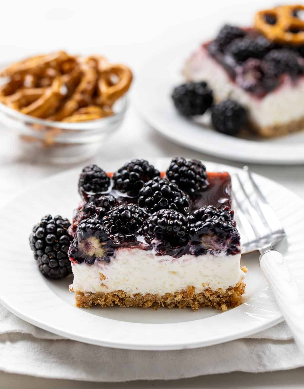 This Blackberry Pretzel Salad is a sweet and salty layered dessert made with a crunchy pretzel crust, creamy cheesecake-style filling, and a glossy black cherry gelatin layer packed with juicy blackberries. It’s creamy, fruity, and perfectly refreshing! White plates with Blackberry Pretzel Salad pieces on them on a white table.
