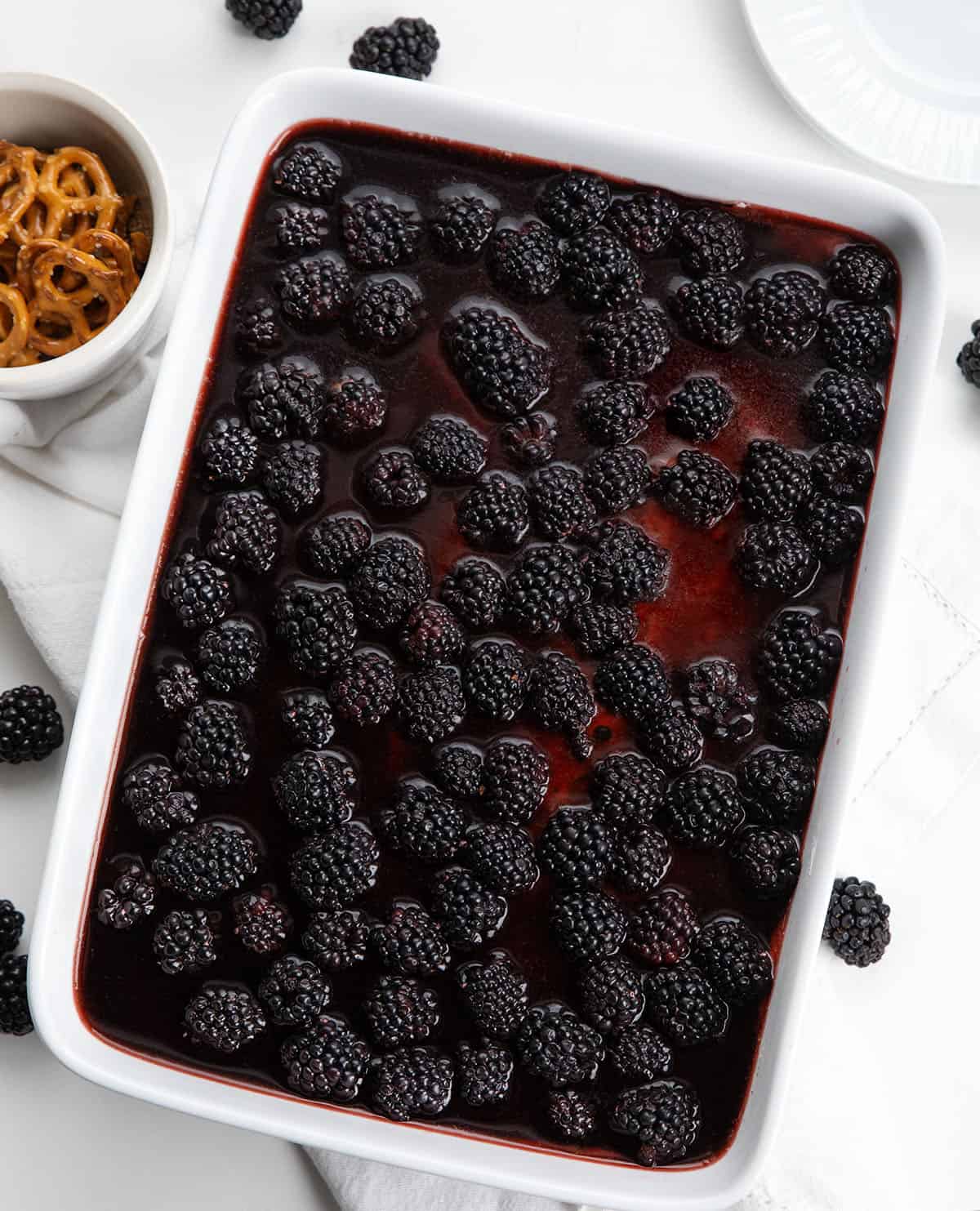 This Blackberry Pretzel Salad is a sweet and salty layered dessert made with a crunchy pretzel crust, creamy cheesecake-style filling, and a glossy black cherry gelatin layer packed with juicy blackberries. It’s creamy, fruity, and perfectly refreshing! Pan of Blackberry Pretzel Salad on a white table from overhead.