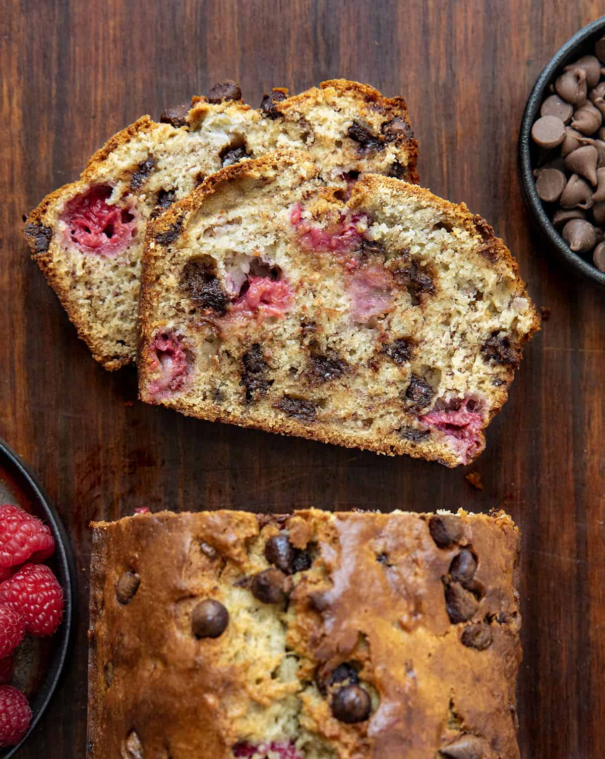 This chocolate raspberry banana bread is soft, moist, and packed with melty chocolate chips and juicy raspberries in every bite. Loaf of Chocolate Raspberry Banana Bread on a wooden table with a couple of pieces of the bread cut and laying flat on the table.