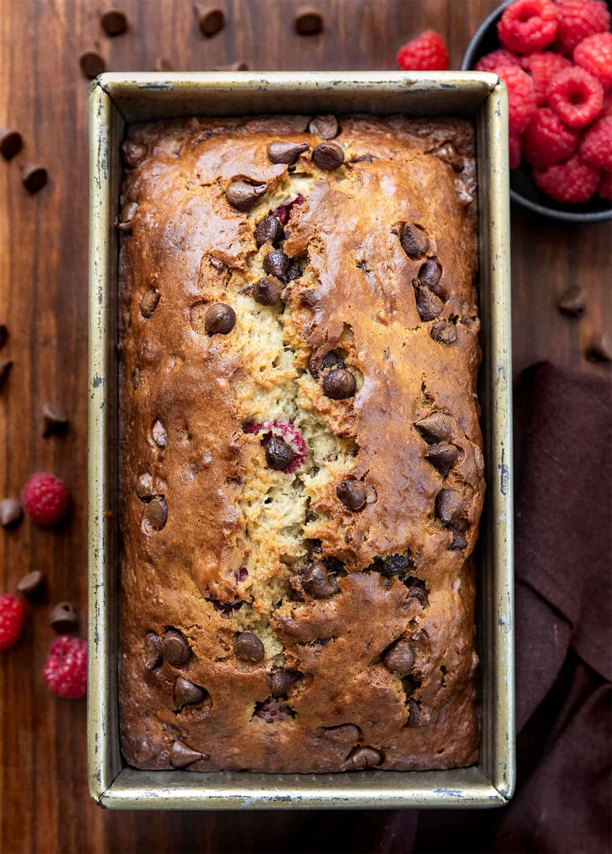 This chocolate raspberry banana bread is soft, moist, and packed with melty chocolate chips and juicy raspberries in every bite. Loaf of Chocolate Raspberry Banana Bread surrounded by raspberries and chocolate from overhead.
