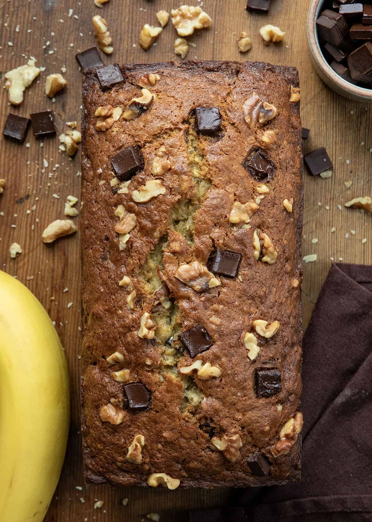 This Chunky Monkey Banana Bread is soft, ultra moist, and loaded with ripe bananas, melty chocolate chunks, and crunchy walnuts in every bite. Itโs a cozy, bakery-style quick bread that leans a little indulgent in the best way! Loaf of Chunky Monkey Banana Bread on a wooden table from overhead.