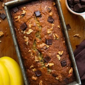 Loaf of Chunky Monkey Banana Bread in the pan on a wooden table surround by bananas and chocolate chunks.
