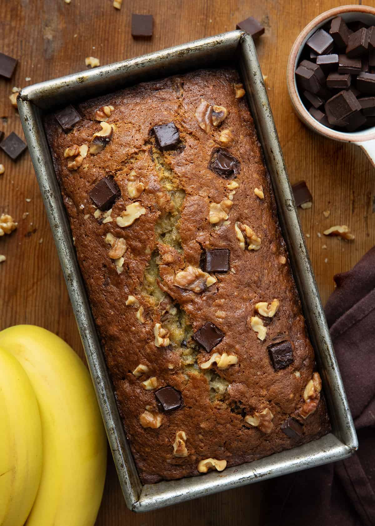 Loaf of Chunky Monkey Banana Bread in the pan on a wooden table surround by bananas and chocolate chunks.