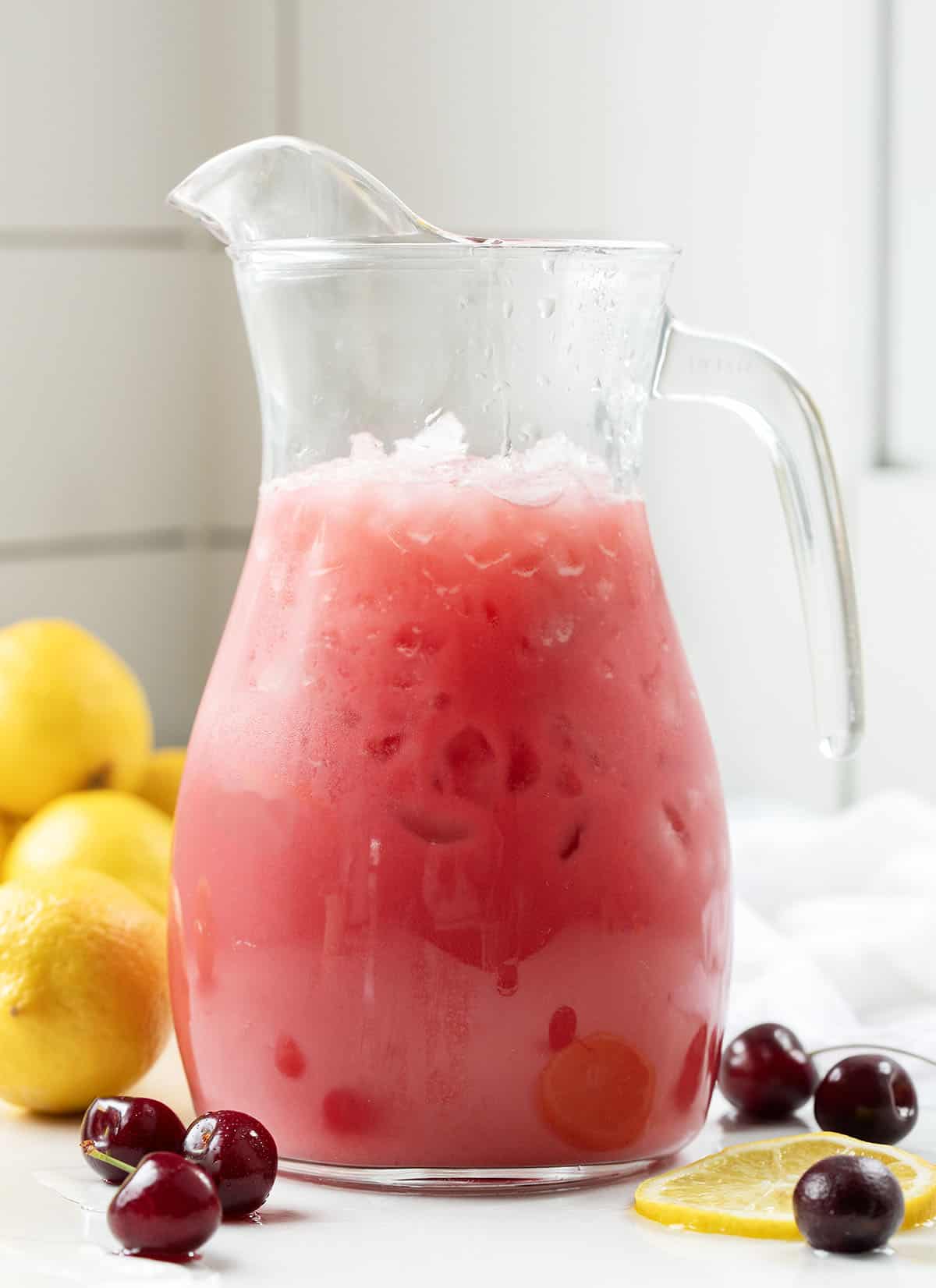 Pitcher of Creamy Cherry Lemonade surrounded by lemons and cherries on a white counter.