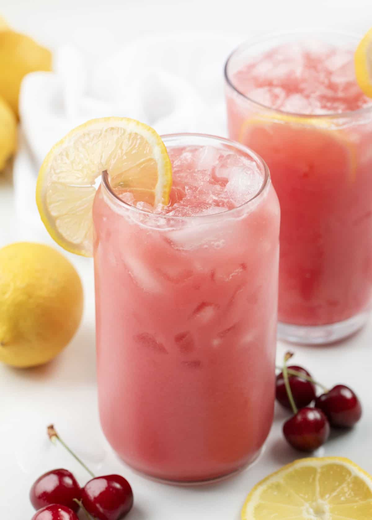 Glasses of Creamy Cherry Lemonade on a white table with cherries and lemons.