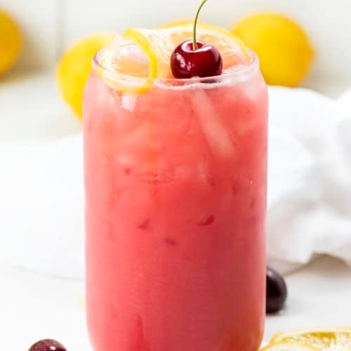 Glass of Creamy Cherry Lemonade on a white table with lemons and cherries.