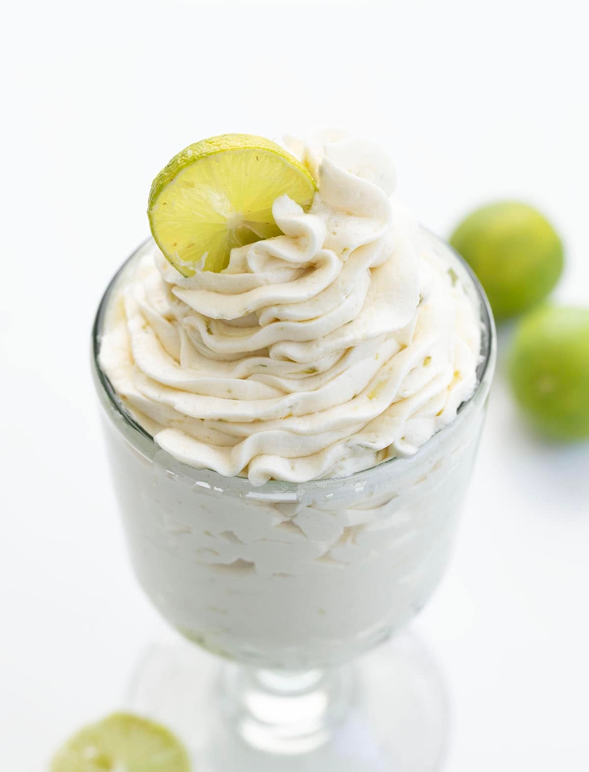 This Key Lime Ermine Frosting is light, silky, and full of bright citrus flavor from fresh key lime juice and zest. It whips up into a smooth, cloud-like frosting thatโs less sweet than traditional buttercream but still rich enough to pipe or spread beautifully! Close up of a Glass of Key Lime Ermine Frosting with fresh key limes.