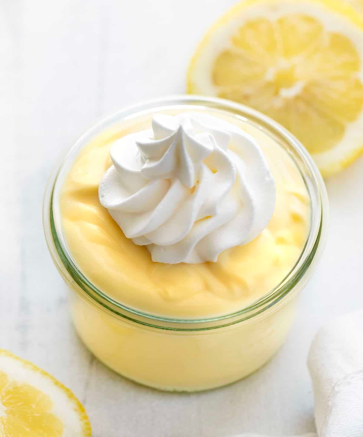 This homemade lemon pudding is smooth, creamy, and full of bright citrus flavor. Made from scratch on the stovetop, it has a rich, silky texture and just the right balance of sweet and tangy. Jae of Lemon Pudding with whipped cream and fresh lemon on a white table.