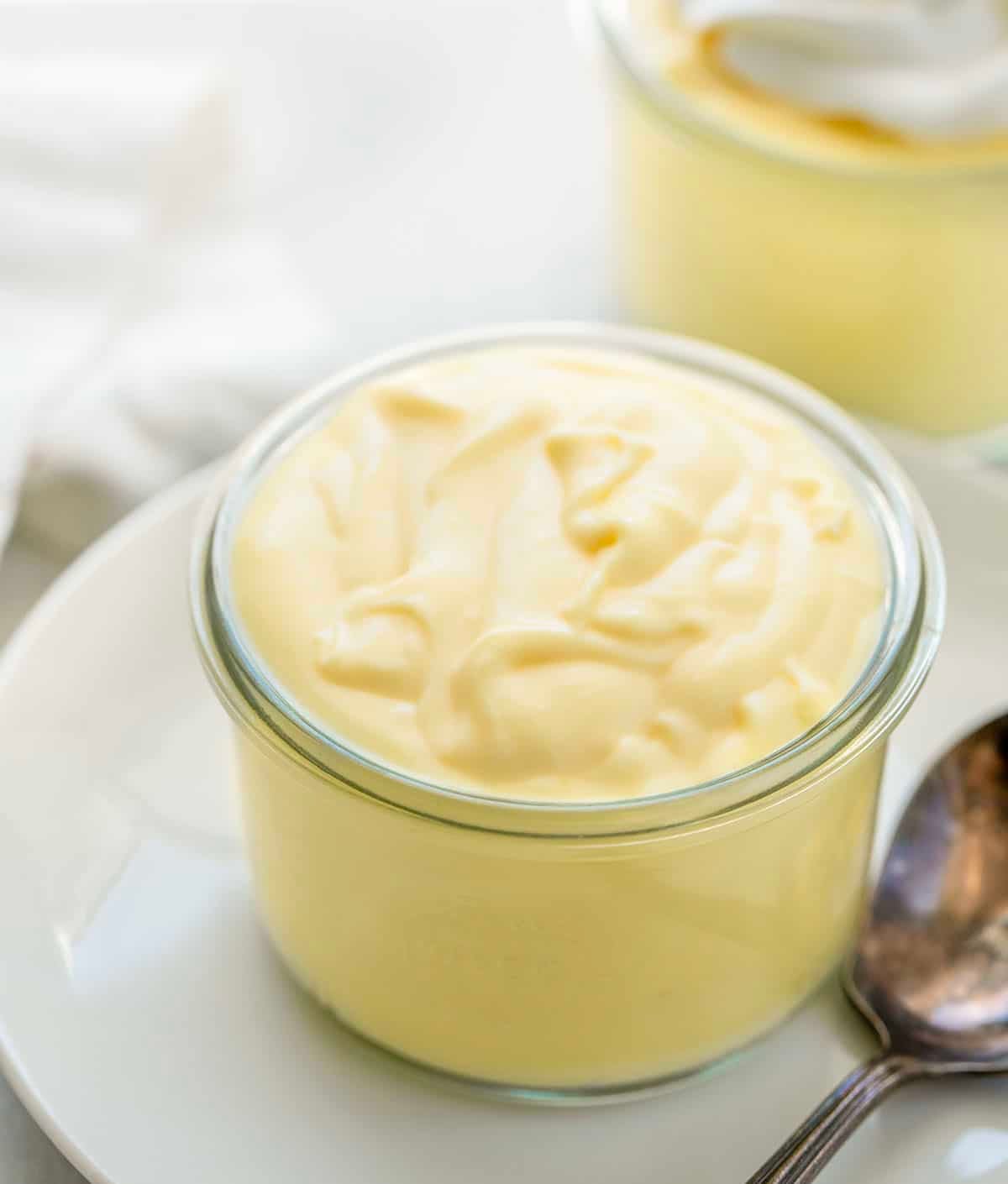 This homemade lemon pudding is smooth, creamy, and full of bright citrus flavor. Made from scratch on the stovetop, it has a rich, silky texture and just the right balance of sweet and tangy. Jar of Lemon Pudding on a white plate with a spoon.