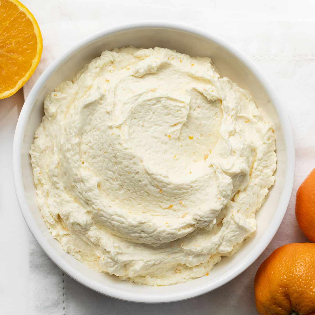 Bowl of Orange Ermine Frosting on a white table with fresh oranges around.