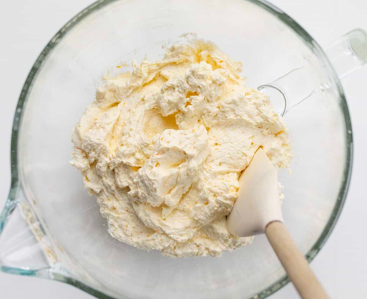 Glass stand mixer bowl filled with Orange Ermine Frosting and a spatula.