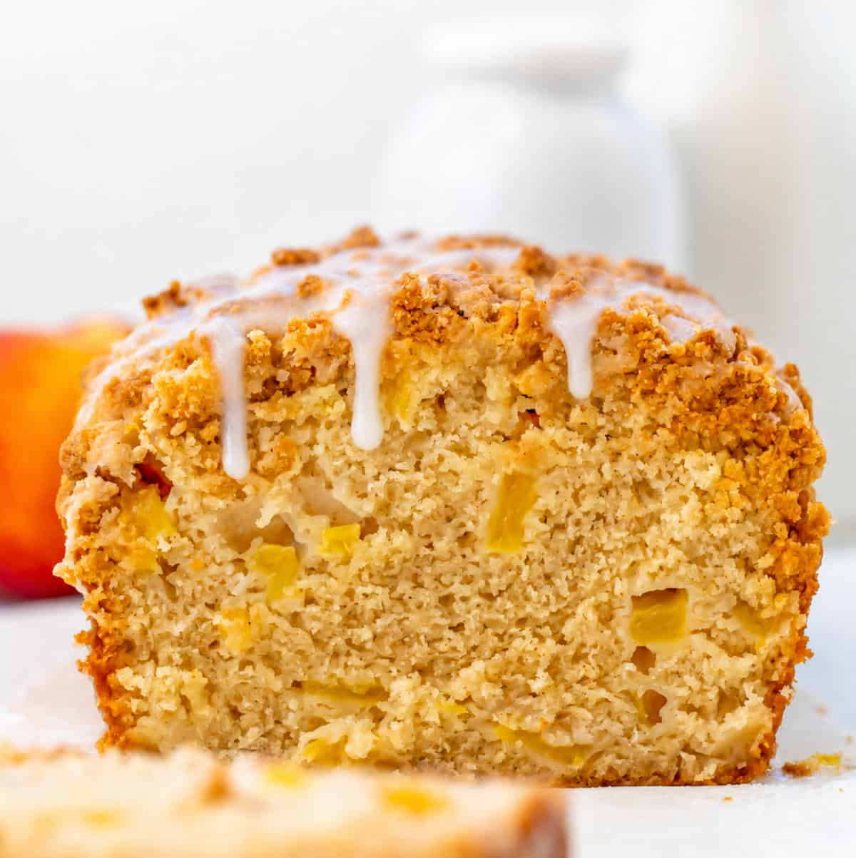 This Peach Cobbler Bread is an easy quick bread that tastes just like peach cobbler in loaf form. It’s soft and tender with juicy peaches in every bite, plus a buttery cinnamon crumble baked right on top! Loaf of Peach Cobbler Bread with glaze drizzling over edge.