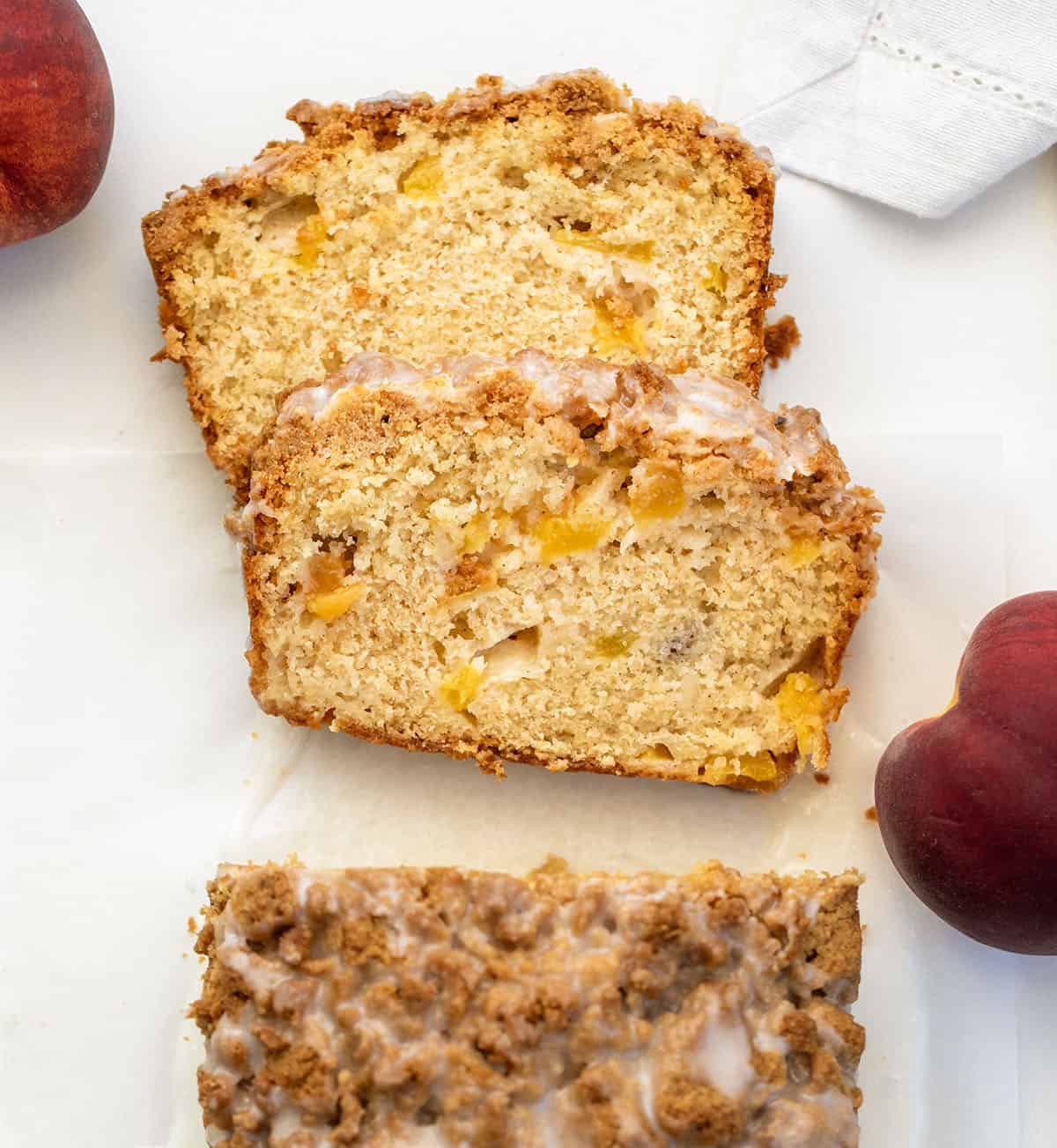 This Peach Cobbler Bread is an easy quick bread that tastes just like peach cobbler in loaf form. It’s soft and tender with juicy peaches in every bite, plus a buttery cinnamon crumble baked right on top! Looking down on a loaf of Peach Cobbler Bread on a white table with a couple of pieces cut and laying flat.