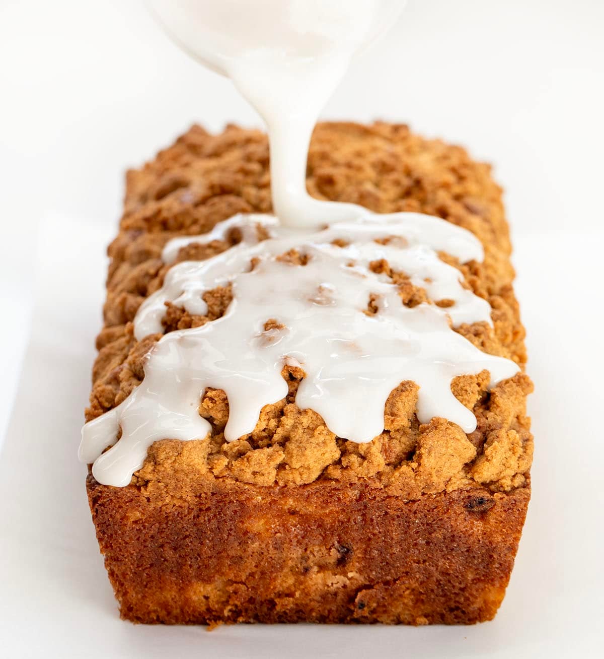 This Peach Cobbler Bread is an easy quick bread that tastes just like peach cobbler in loaf form. It’s soft and tender with juicy peaches in every bite, plus a buttery cinnamon crumble baked right on top! Drizzling glaze over Peach Cobbler Bread on a white table.
