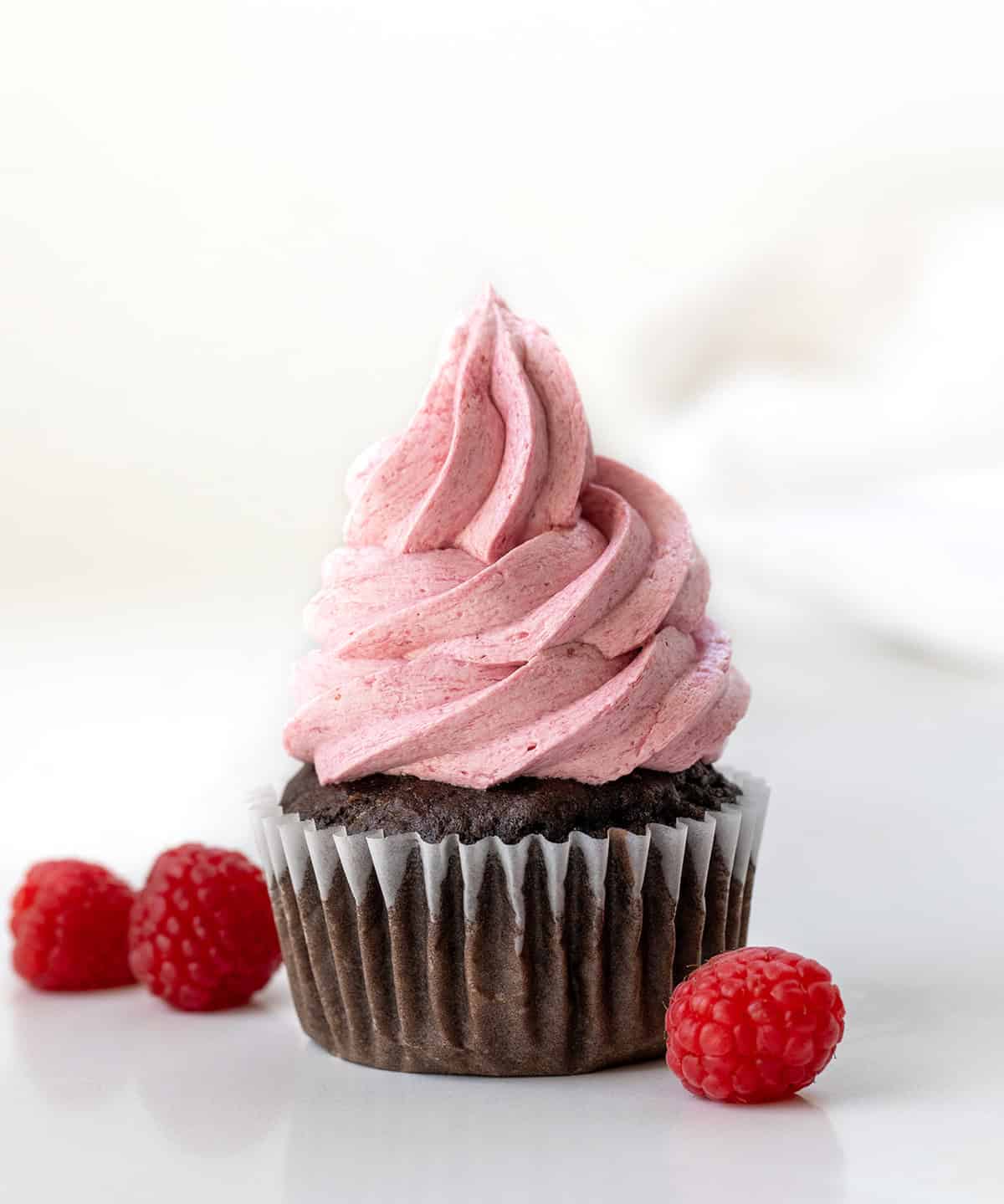 Chocolate cupcake with Raspberry Ermine Frosting piped on top and surrounded by raspberries on a white table.