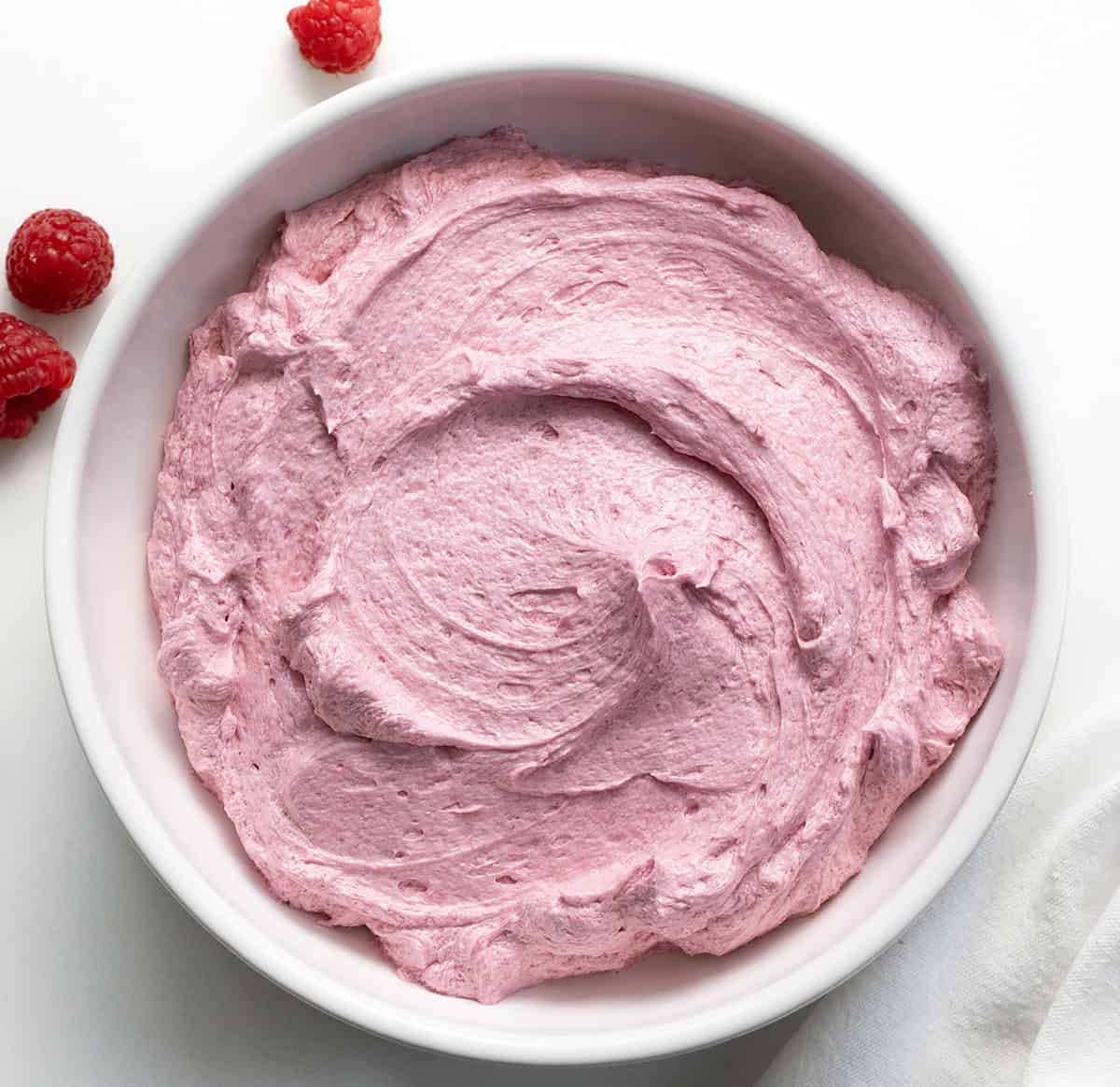 White bowl filled with Raspberry Ermine Frosting on a white table with a few fresh raspberries.