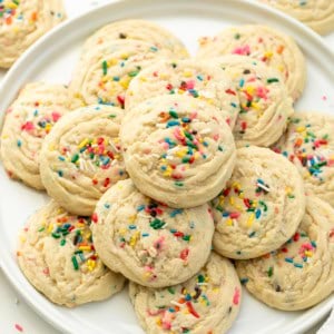 White platter with Sprinkles Sugar Cookies stacked on it on a white table surrounded by more cookies.
