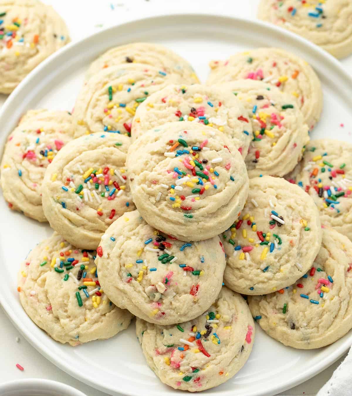 White platter with Sprinkles Sugar Cookies stacked on it on a white table surrounded by more cookies.