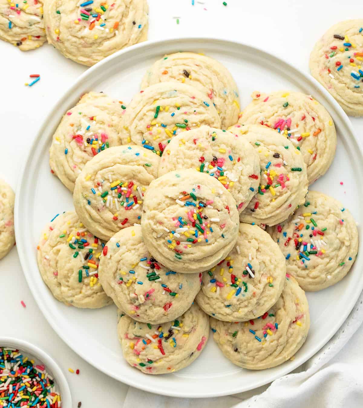 White platter with Sprinkles Sugar Cookies stacked on it on a white table surrounded by more cookies from overhead.