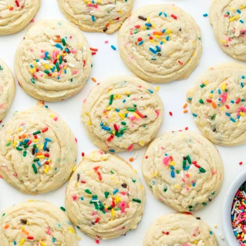Sprinkles Sugar Cookies on a white table from overhead with a bowl of rainbow sprinkles nearby.