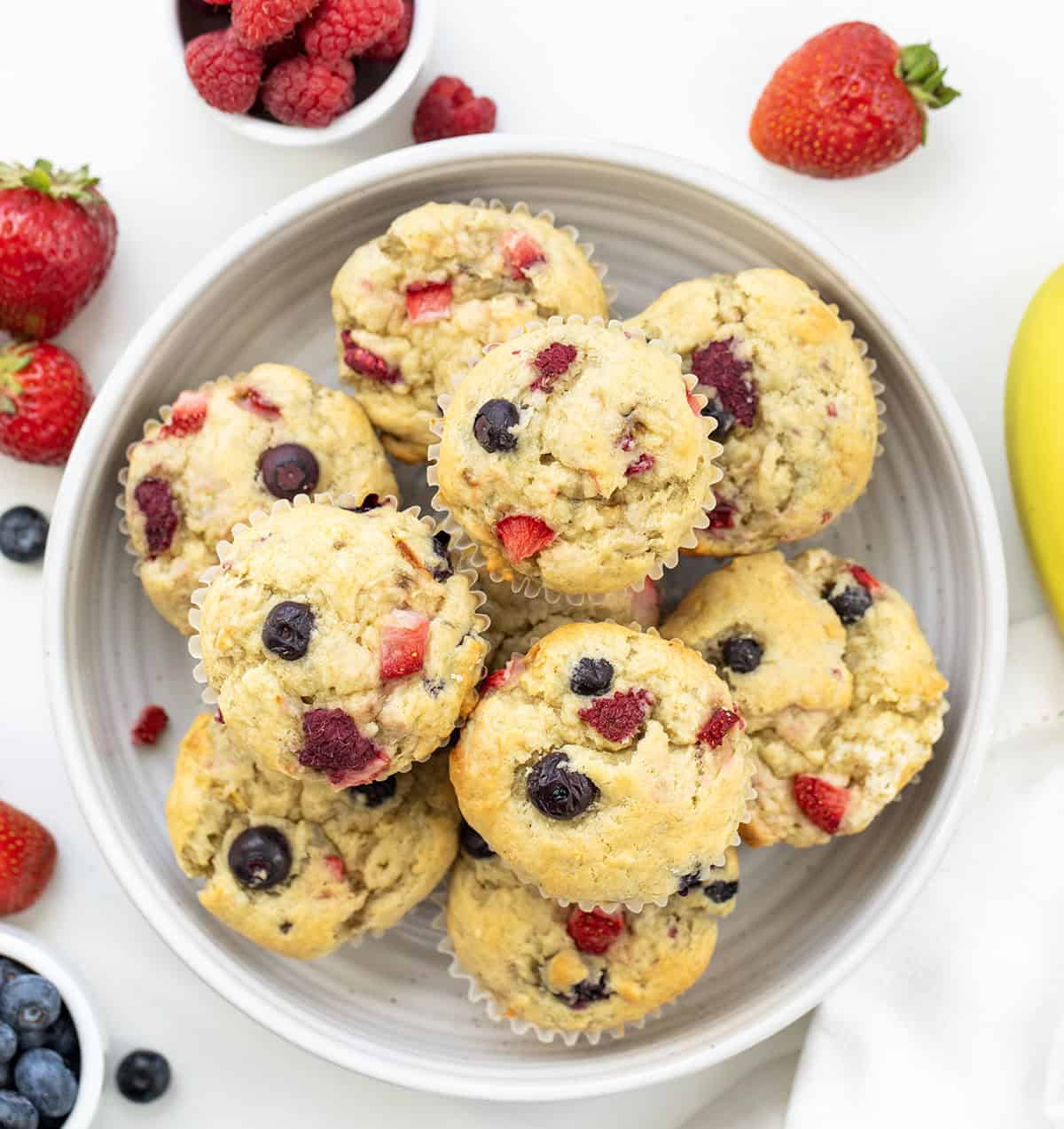 White bowl of Triple Berry Banana Muffins on a white table with fresh raspberries, strawberries, blueberries, and banana.