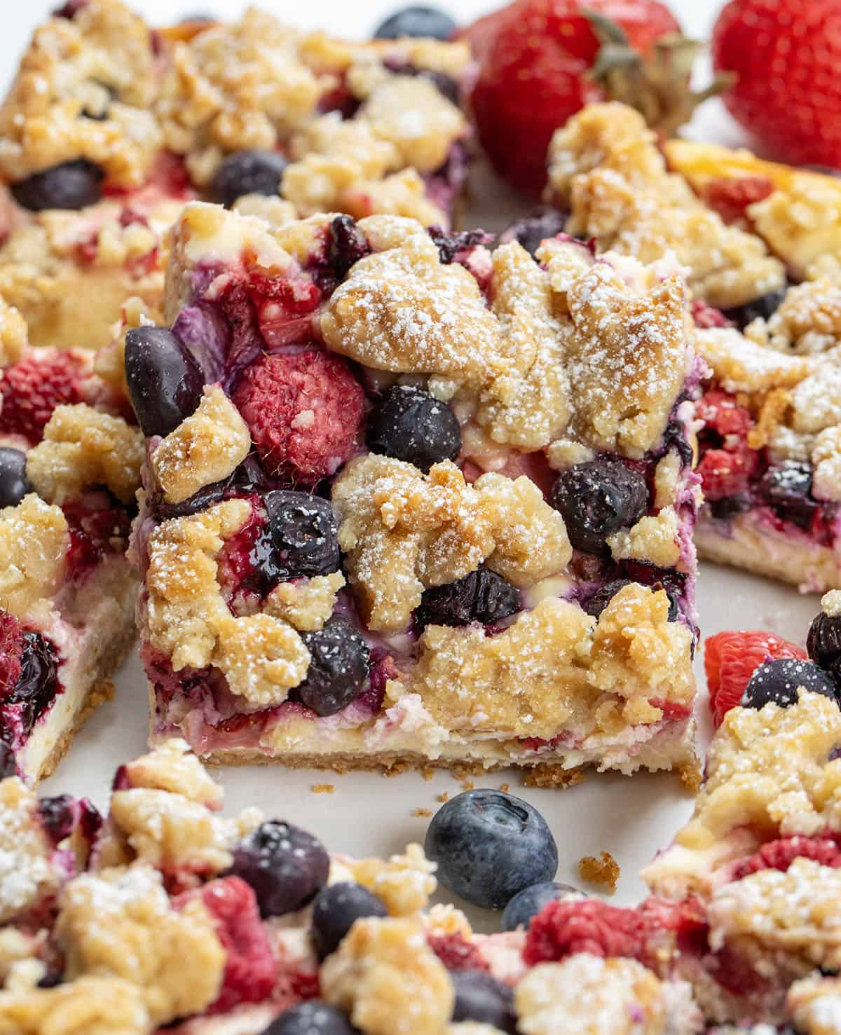 These Triple Berry Cheesecake Bars are a simple,crowd-pleasing dessert made with a buttery graham cracker crust, creamy cheesecake filling, and a mix of fresh blueberries, strawberries, and raspberries! Close up of a Triple Berry Cheesecake Bar.