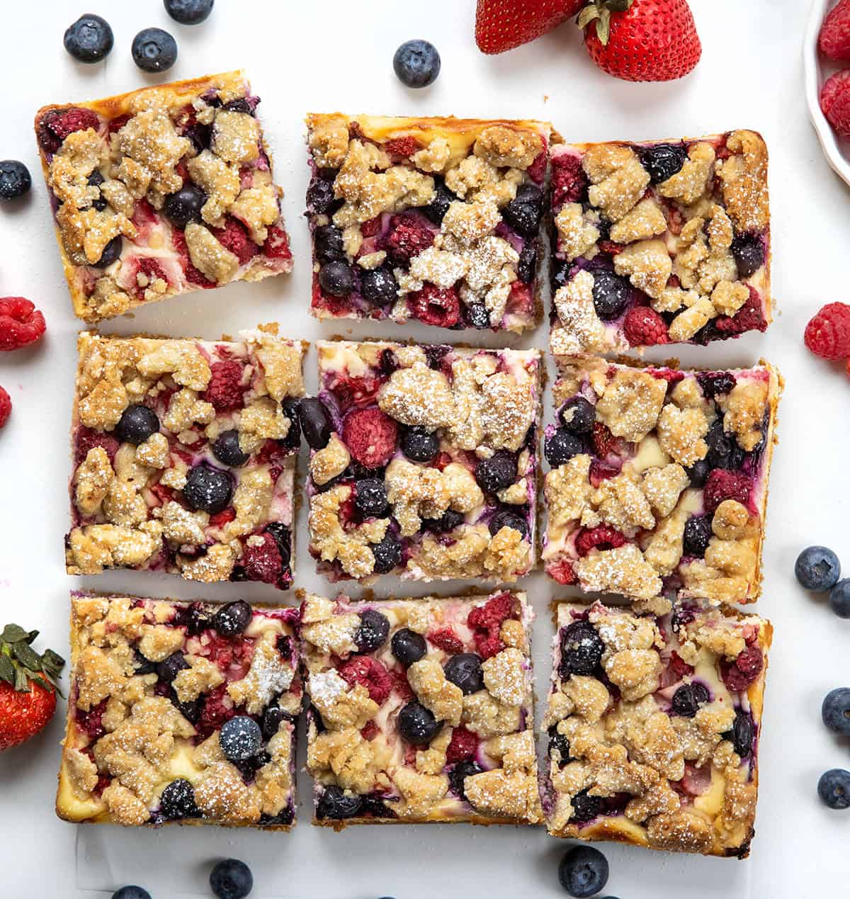 These Triple Berry Cheesecake Bars are a simple,crowd-pleasing dessert made with a buttery graham cracker crust, creamy cheesecake filling, and a mix of fresh blueberries, strawberries, and raspberries! Triple Berry Cheesecake Bars on a white table cut into squares and surrounded by fresh fruit.
