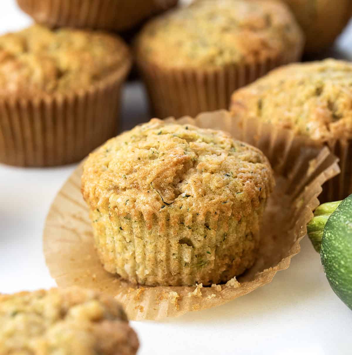 Unwrapped Zucchini Muffin on a white table with zucchini.