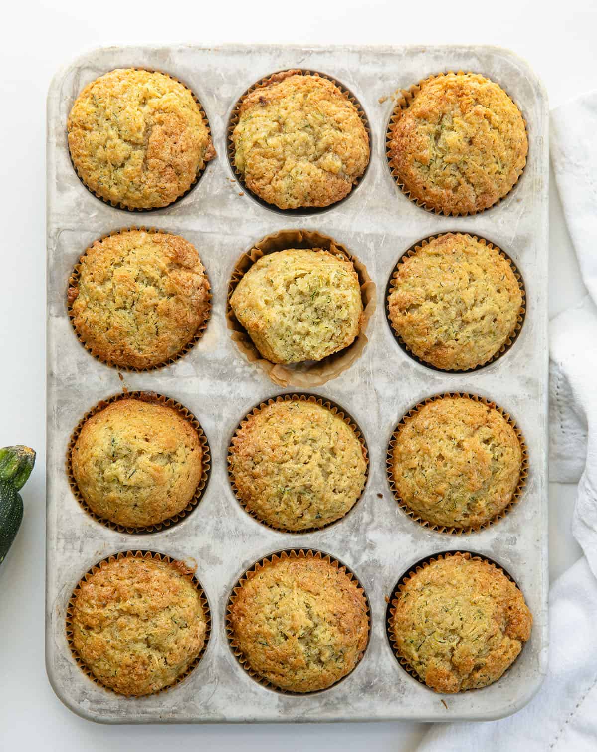 Zucchini Muffins in a muffin pan with one muffin halved showing the inside texture.