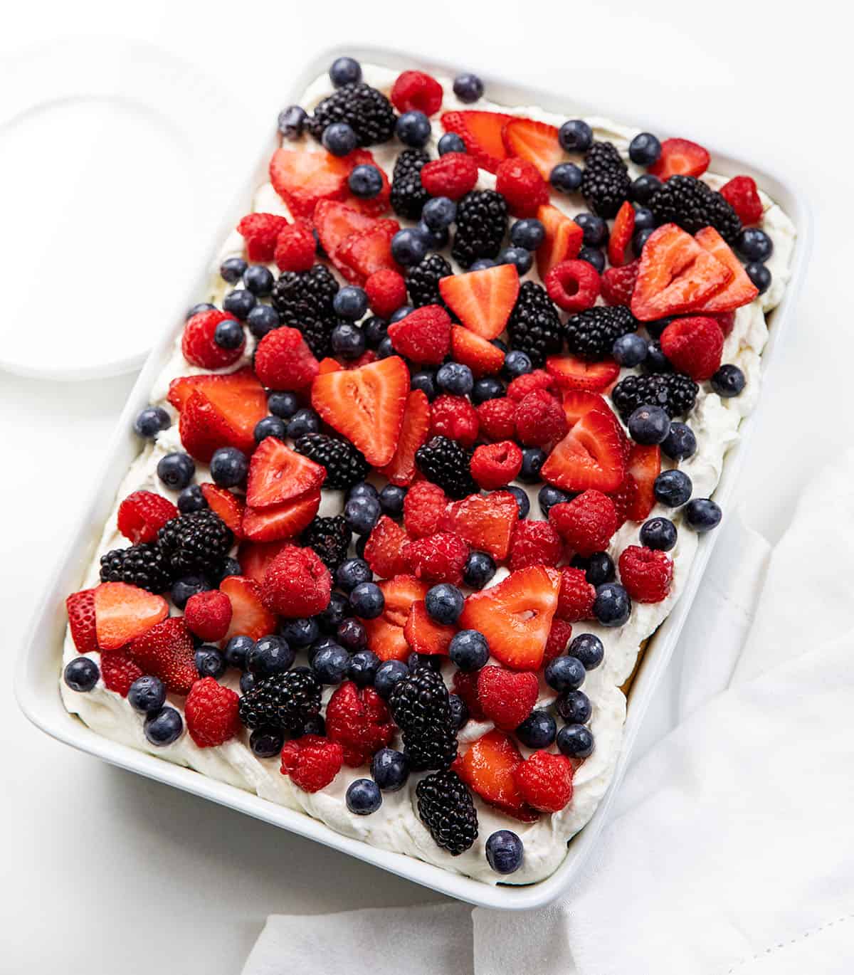 Berries & Cream Sheet Cake on a white table with white plates and a white hand towel next to the cake pan.