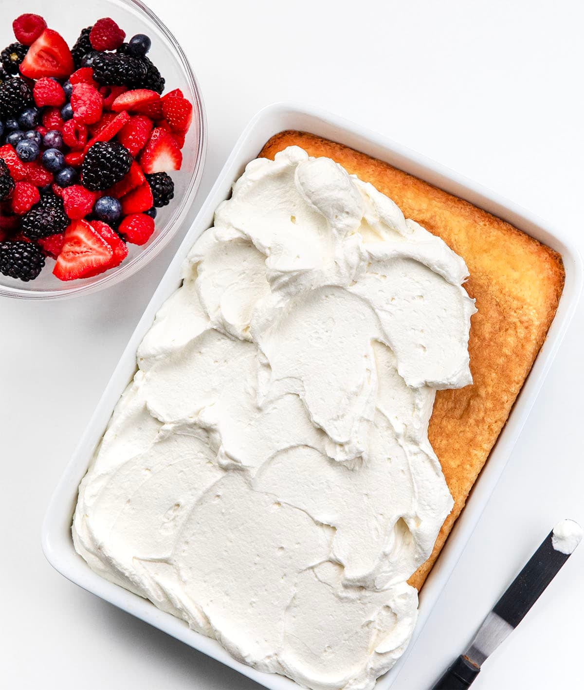Spreading stabilized whipped cream over the best white sheet cake and a bowl of fresh berries next to the cake pan.