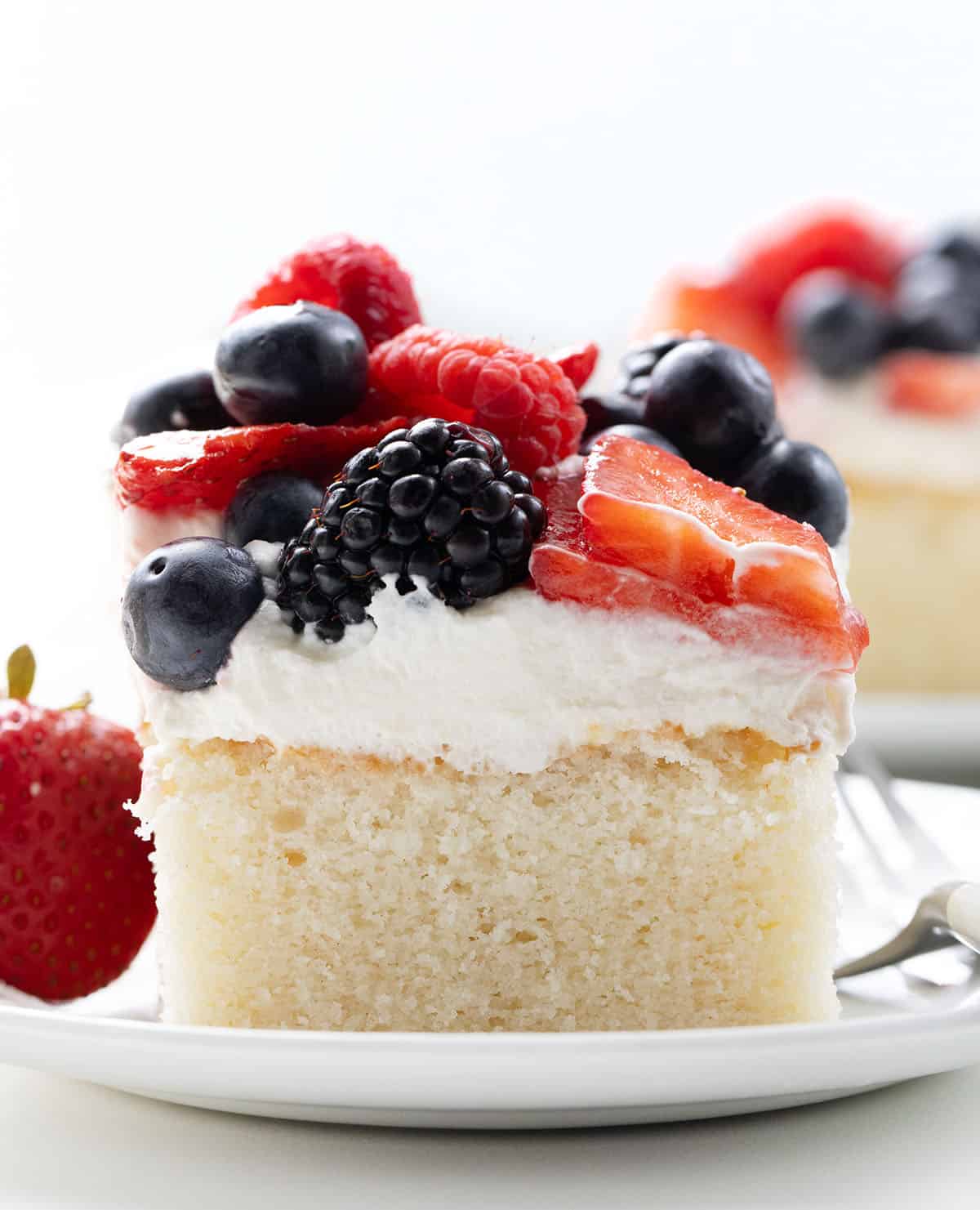 Close up of a piece of Berries & Cream Sheet Cake on a white plate with another piece in the background.