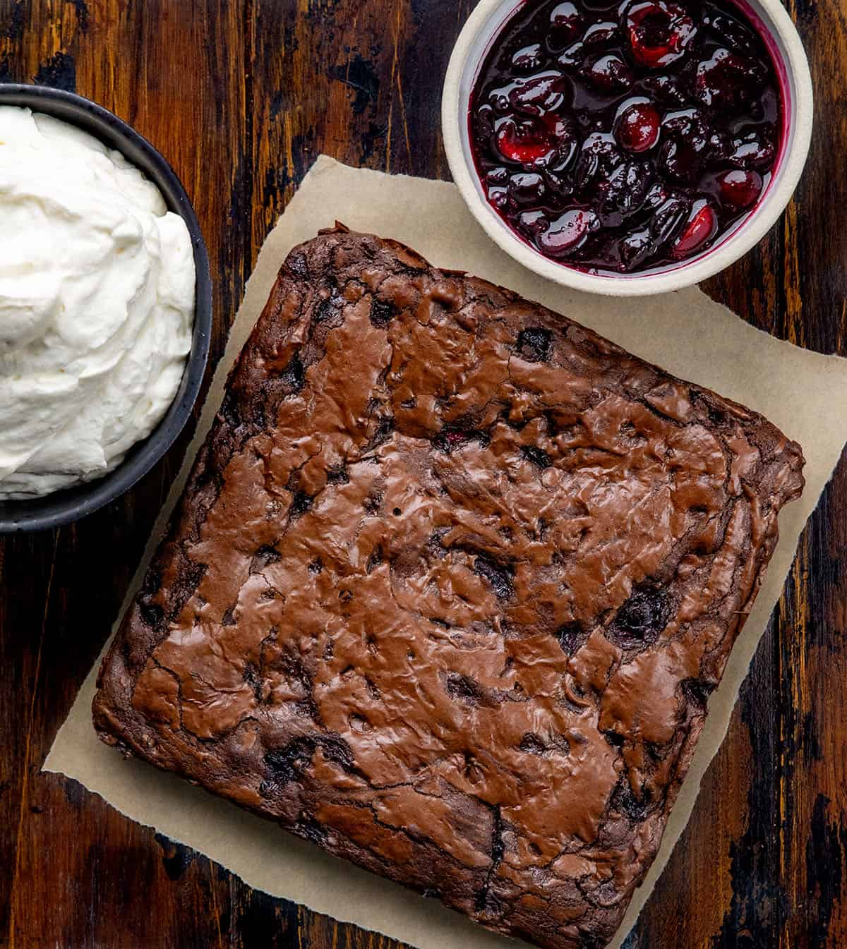 Cherry brownies, stabilized whipped cream, and cherry sauce on a wooden table from overhead.