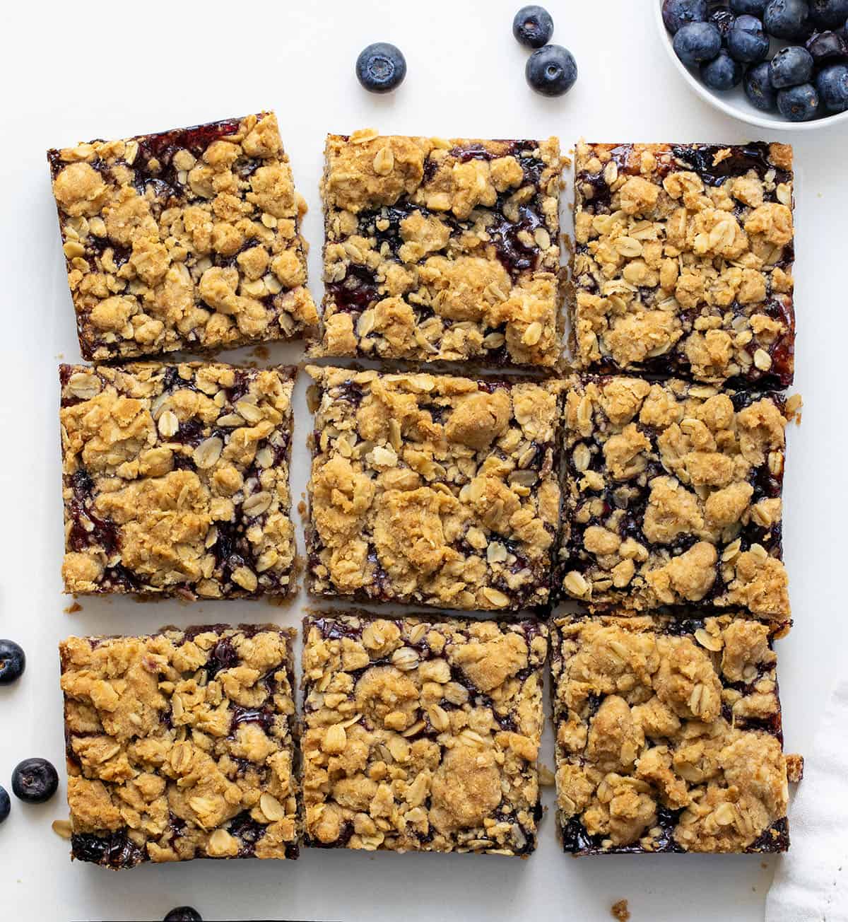 Blueberry Oatmeal Crumble Bars with buttery oat crust, blueberry filling, and golden crumble topping. Blueberry Oatmeal Crumble Bars cut into squares on a white table from overhead.