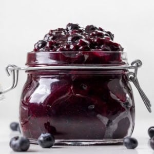 Jar of Blueberry Pie Filling on a white table with fresh blueberries.