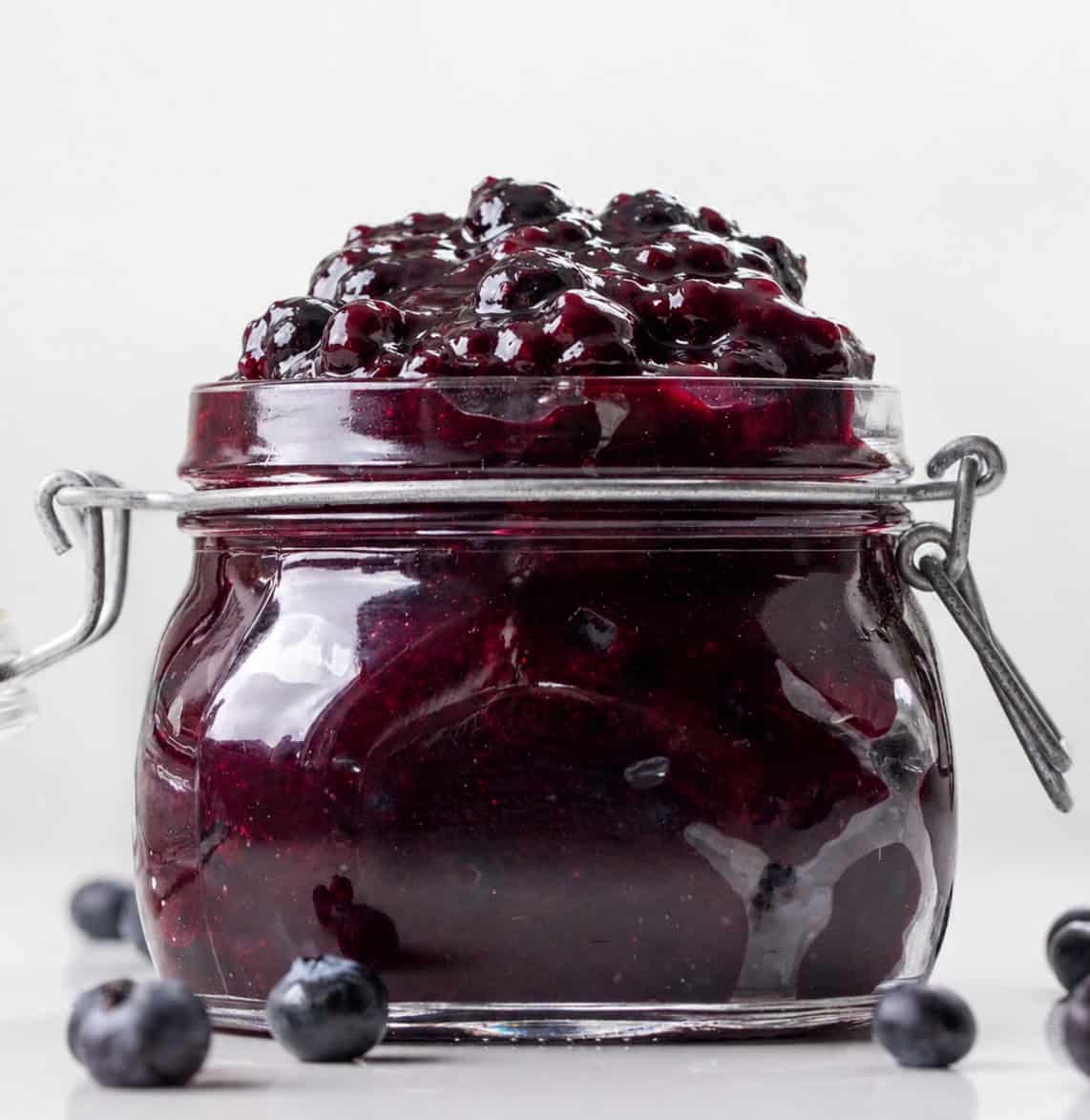 Jar of Blueberry Pie Filling on a white table with fresh blueberries.