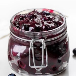 Jar of Blueberry Pie Filling on a white table with fresh blueberries.