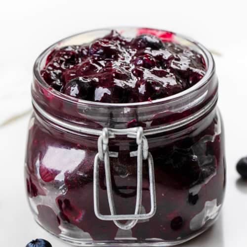 Jar of Blueberry Pie Filling on a white table with fresh blueberries.