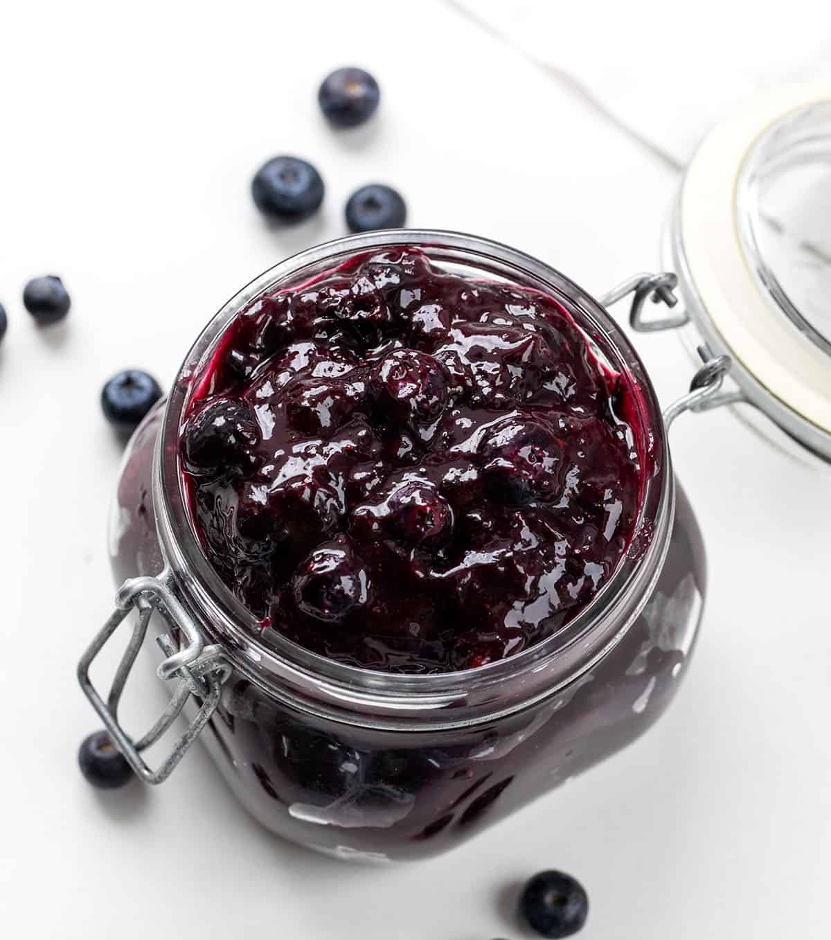 Jar of Blueberry Pie Filling on a white table with fresh blueberries.