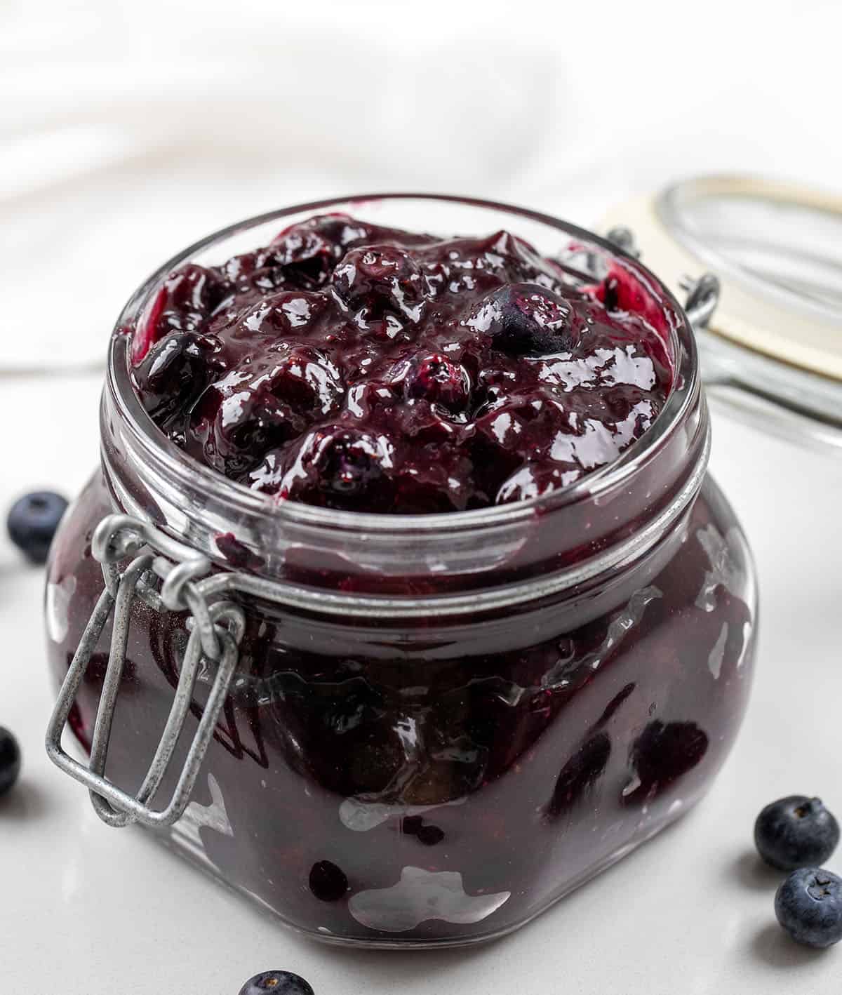 Jar of Blueberry Pie Filling on a white table with fresh blueberries.
