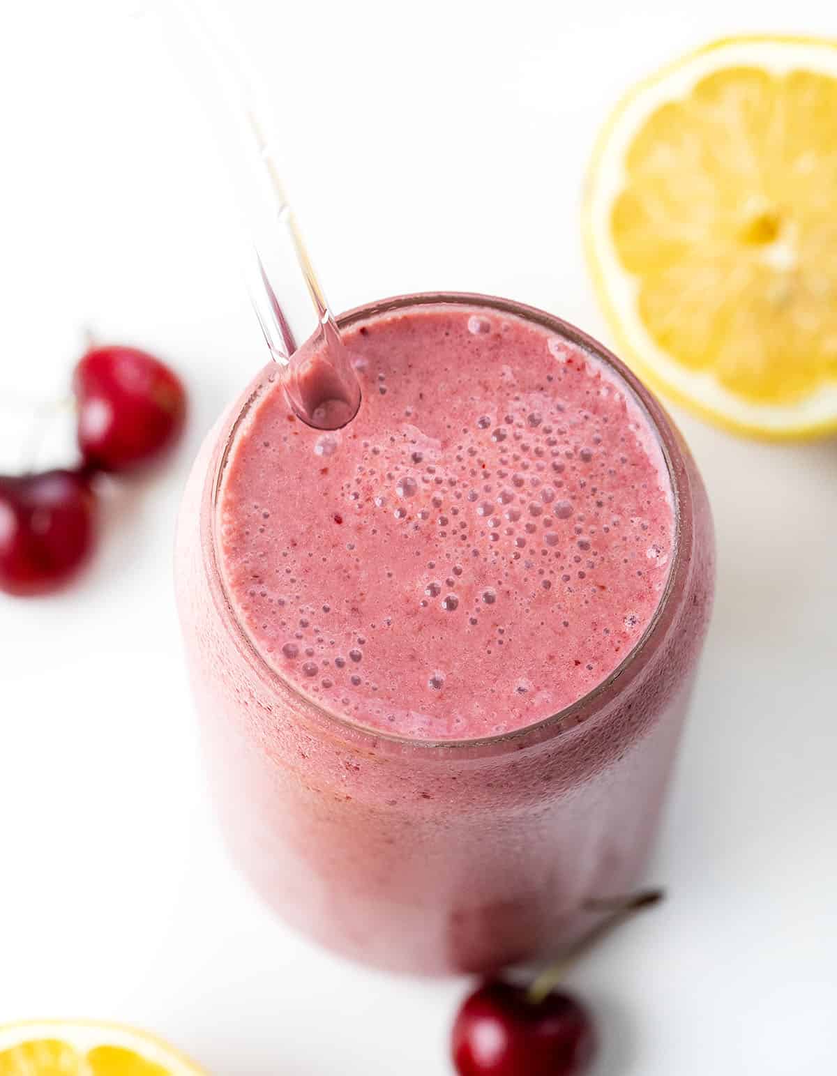 Looking down on a glass of Cherry Frosted Lemonade on a white table with fresh cherries and lemon.