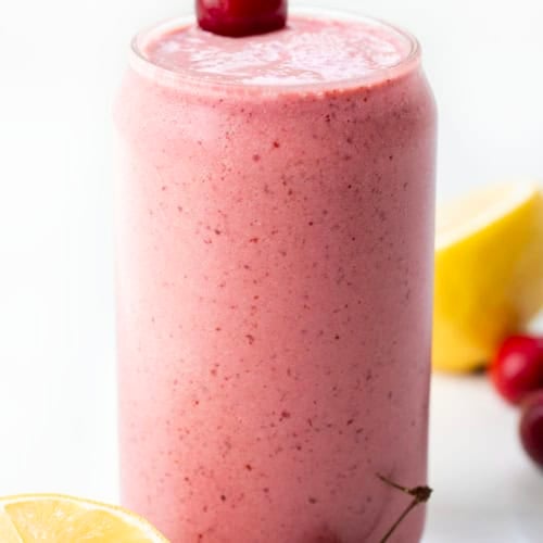 Glass of Cherry Frosted Lemonade on a white table with lemon and cherries.