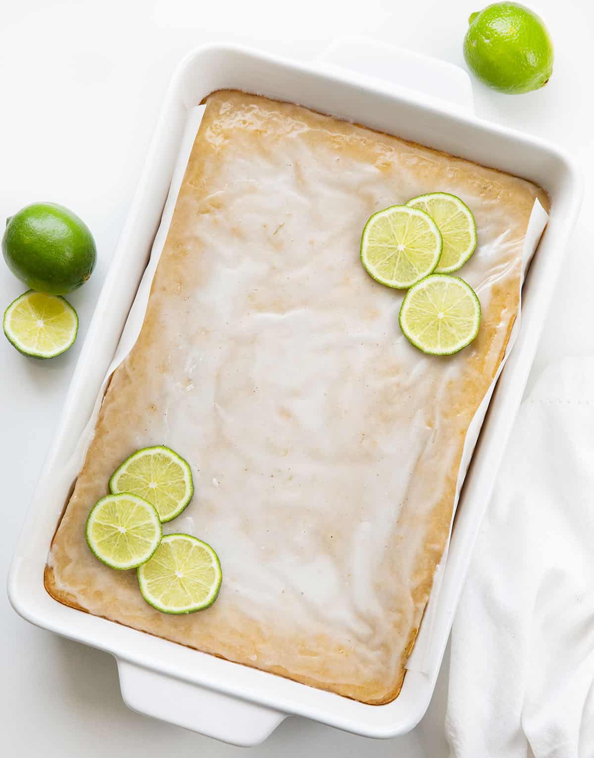 These Lime Brownies are soft, chewy, and bursting with fresh citrus flavor in every bite. Made with brown sugar for a blondie-like texture and finished with a sweet-tart lime glaze, theyโre the perfect balance of rich and refreshing! White pan filled with lime brownies and fresh lime slices on a white table from overhead.