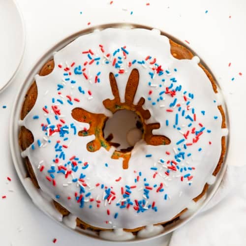 Whole Red, White, and Blue Pound Cake covered in vanilla glaze on a white cake plate on a white table from overhead.