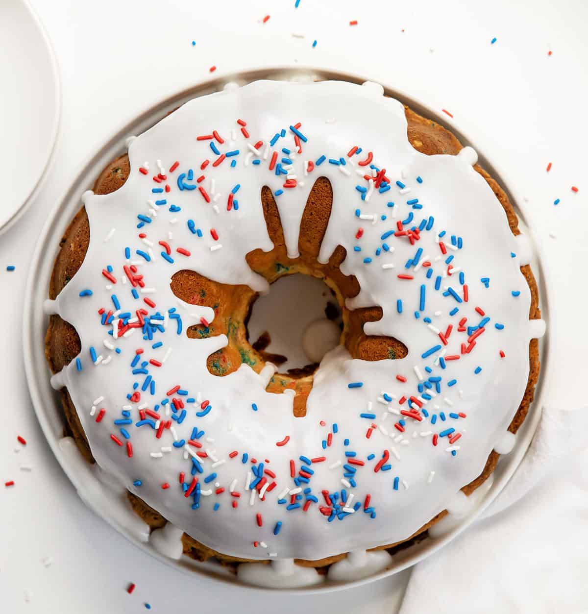 Whole Red, White, and Blue Pound Cake covered in vanilla glaze on a white cake plate on a white table from overhead.
