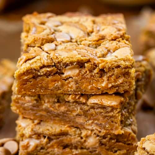 Stack of Peanut Butter Overload Blondies on a wooden table surrounded by more bars and peanut butter morsels.
